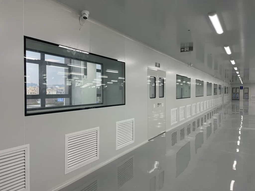 Panoramic view of a clean - area corridor in a hospital. Multiple airtight automatic doors with observation windows are distributed on the walls on both sides. A linear lighting system is installed on the ceiling. The floor is a smooth and reflective antibacterial floor tile, cooperating with the wall air vents to achieve air circulation control.