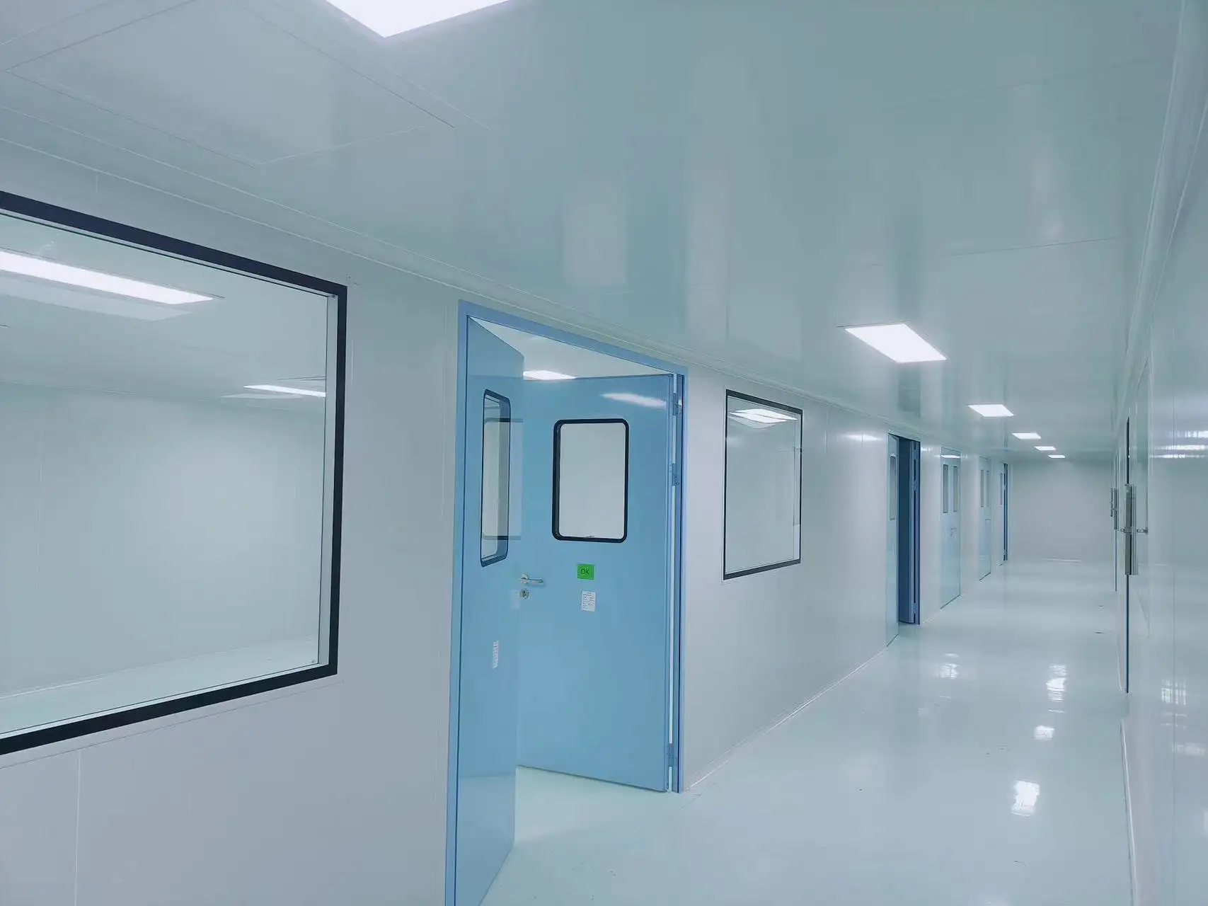 Airtight door in a cleanroom corridor (light blue finish, transparent vision panels), demonstrating particle control and pressure stability in positive-pressure environments (e.g., pharmaceutical, semiconductor facilities).