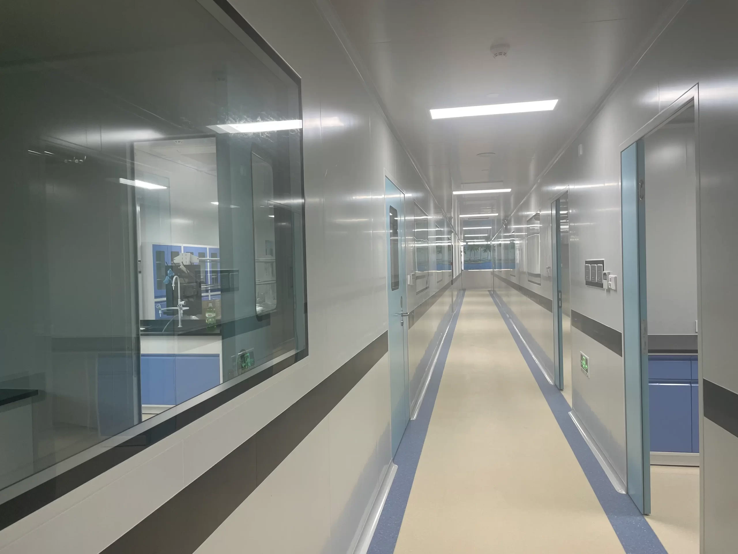 Long corridor of ISO 7 cleanroom with white modular walls, glass windows revealing laboratory interiors, light-colored epoxy flooring with blue dividing lines, and ceiling-mounted LED lighting.