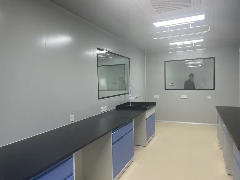 Interior view of ISO 7 cleanroom featuring gray seamless walls, black epoxy workbenches with blue cabinets, ceiling-mounted HEPA filtration unit, and glass viewing windows.