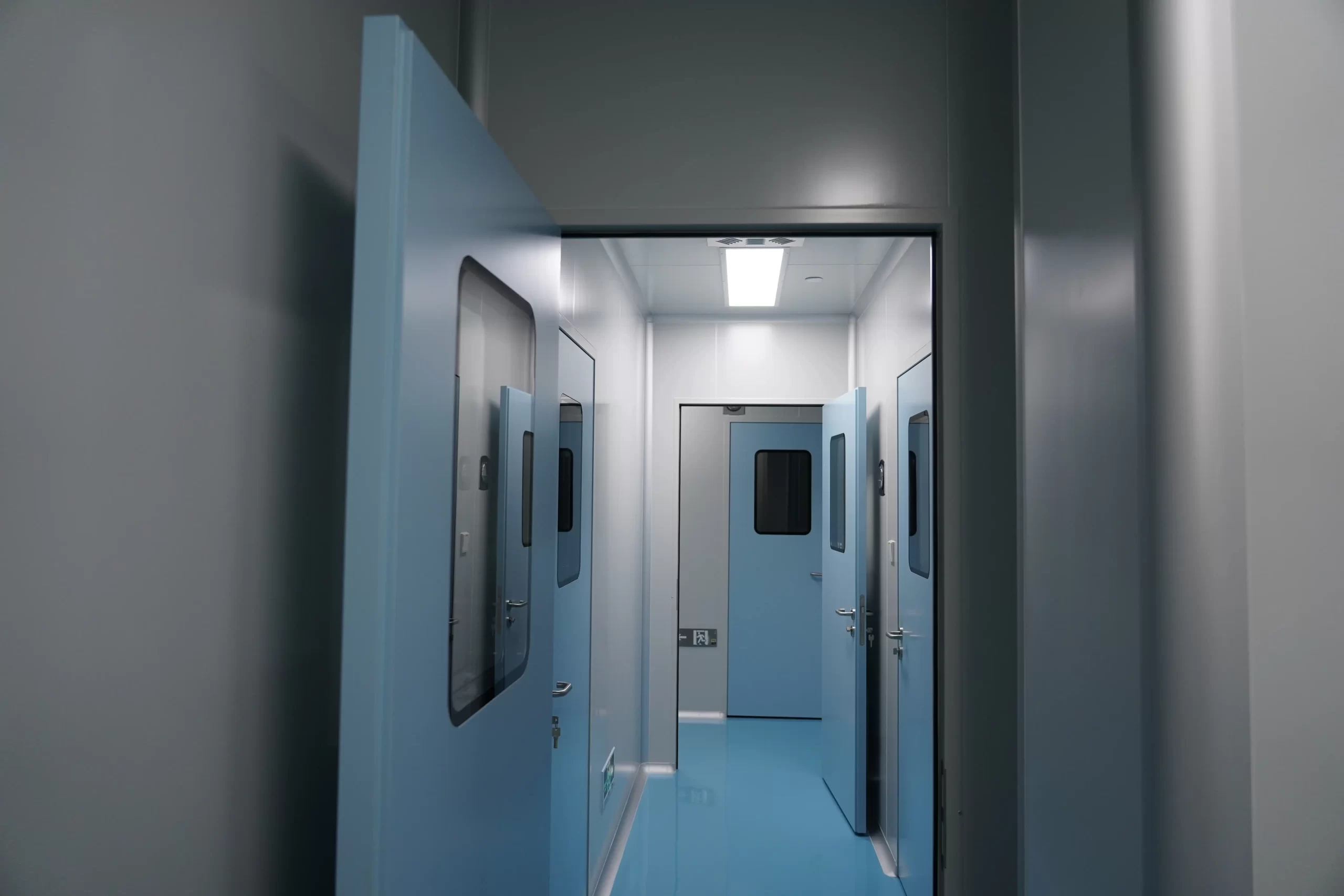 A corridor in a pharmaceutical laboratory with multiple stainless steel cleanroom doors, each with observation windows, maintaining airtightness and hygiene standards for sterile processes.