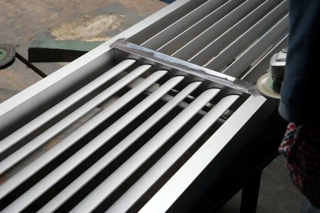 Close-up of a worker polishing an aluminum HVAC grille frame with a power sander, showing parallel slats and a workbench with tools in the background.