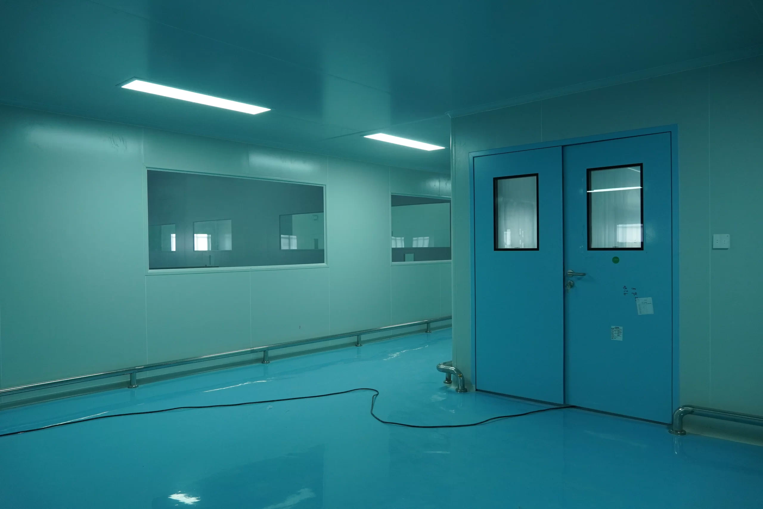 A clean room interior with light blue walls, epoxy floor, and a double-leaf fire door with viewing windows, alongside ceiling lights and wall-mounted HVAC piping.