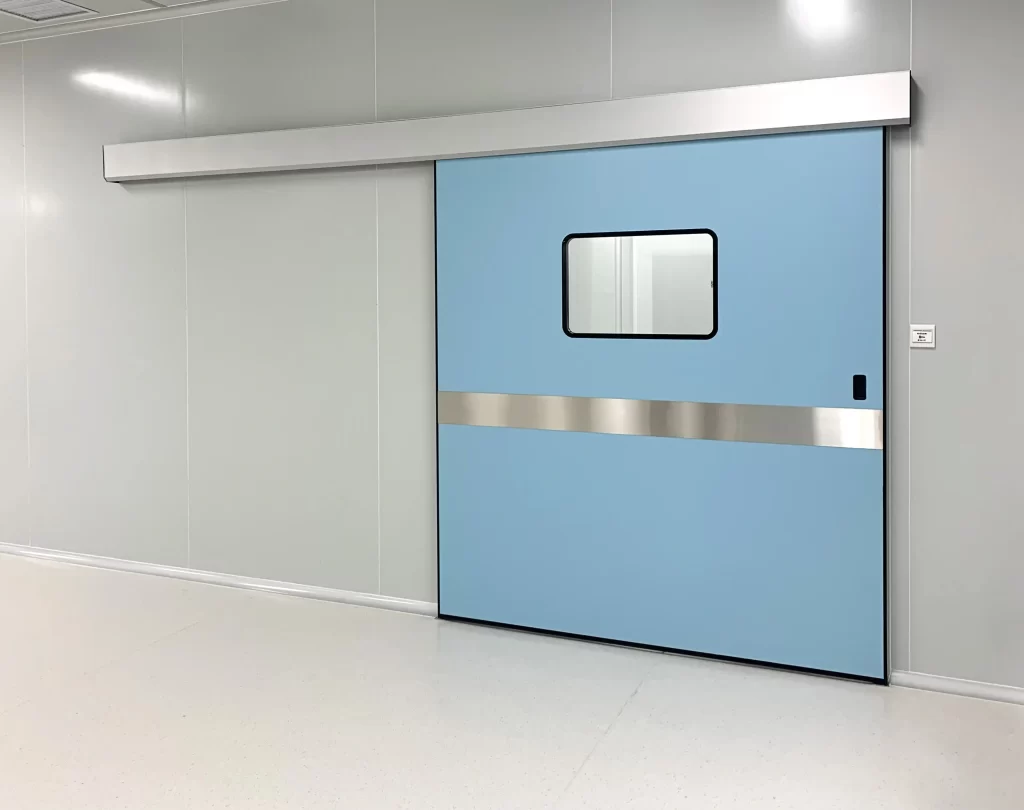 Light blue automatic sliding door in a cleanroom, with a small rectangular observation window and horizontal stainless steel strip, set against light gray panel walls and a smooth white floor.