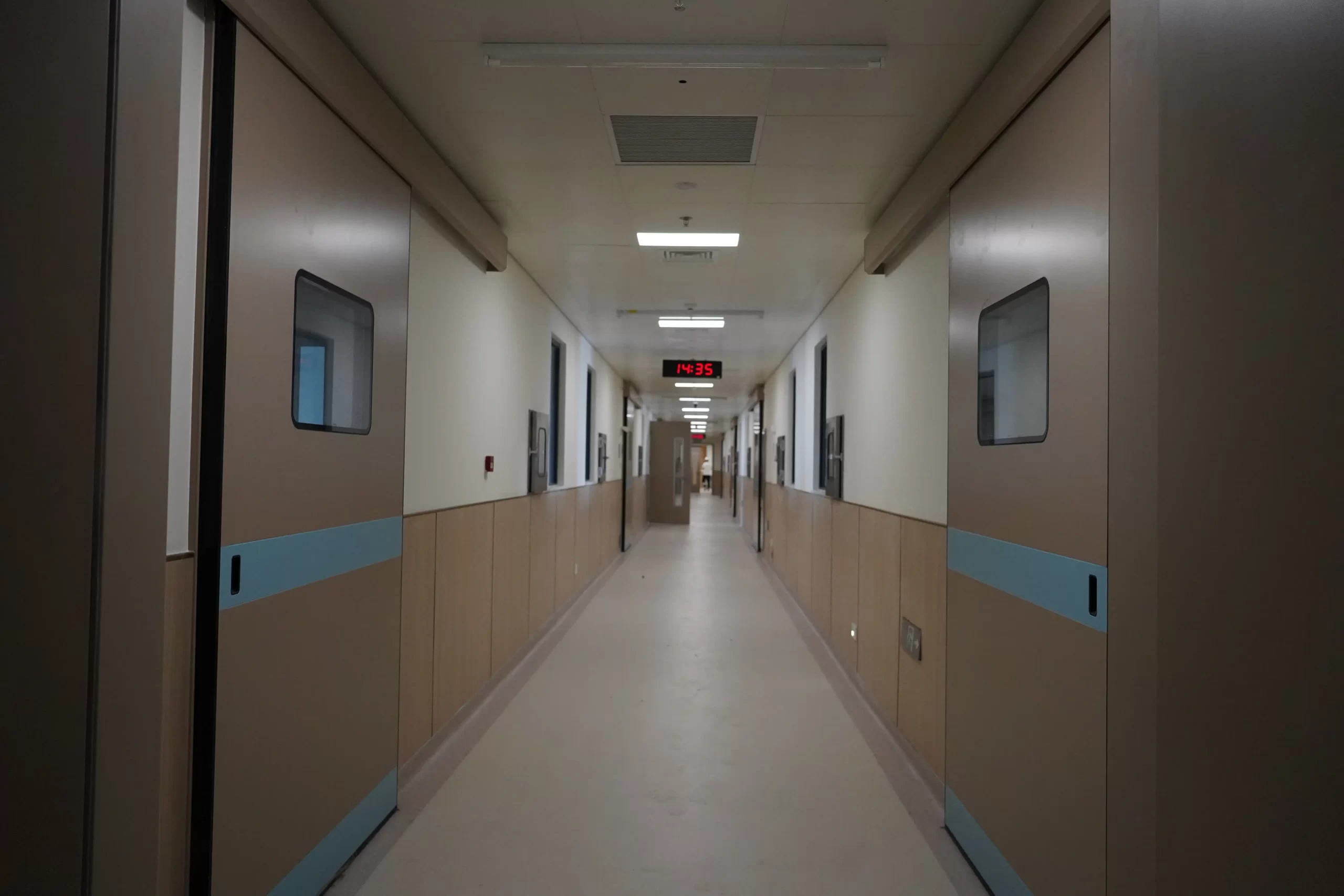 A long hospital corridor with automatic sliding doors on both sides, clean walls, and a digital clock showing 14:35, typical of a medical facility environment.