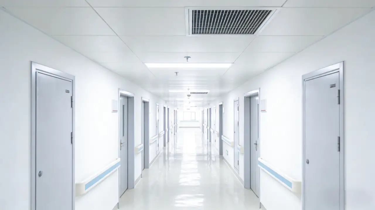 Bright, sterile hospital corridor with white walls, glossy flooring, closed doors, handrails, and a suspended ceiling HVAC diffuser, illuminated by linear LED lights.