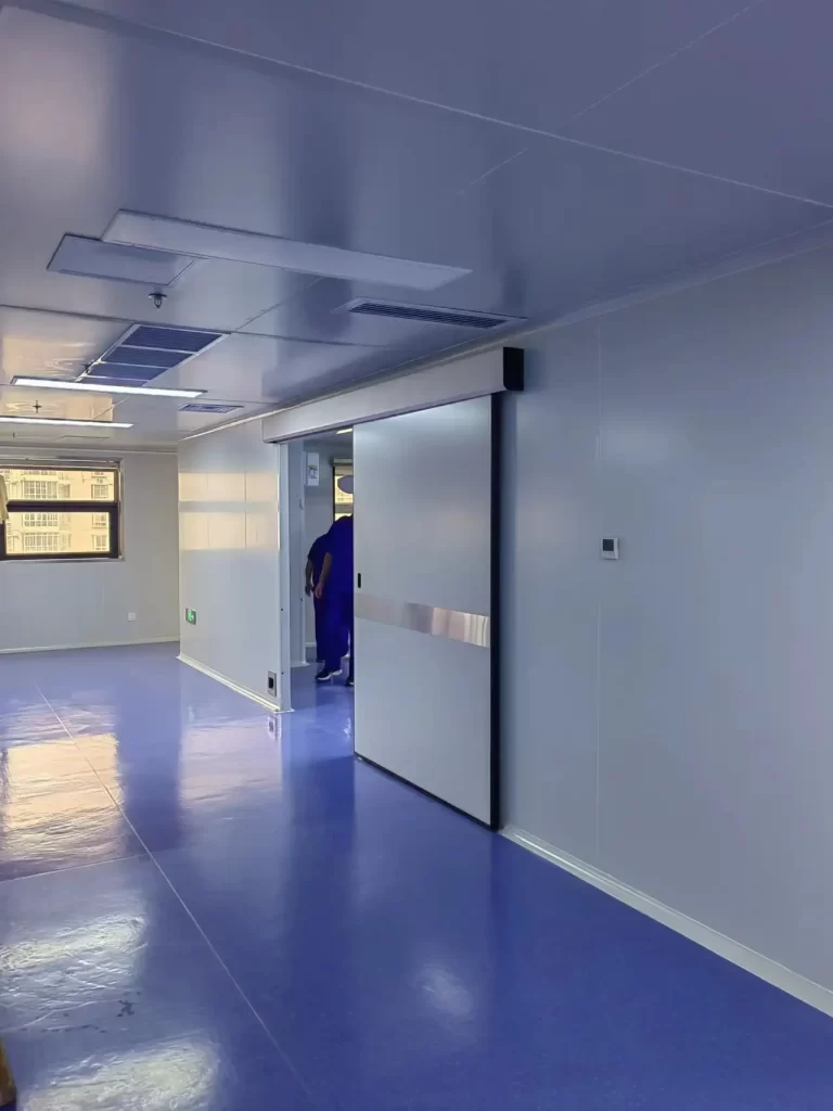 On-site automatic sliding cleanroom door in a pharmaceutical cleanroom, with stainless steel trim, white clean panels, blue epoxy floor, and staff in blue PPE at the doorway.