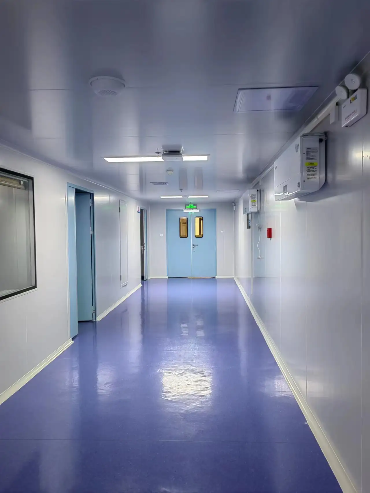 Manual double doors in a cleanroom corridor, with blue epoxy floor, white clean panels, emergency exit sign, and fire safety equipment, showing manual door application in buffer zones.
