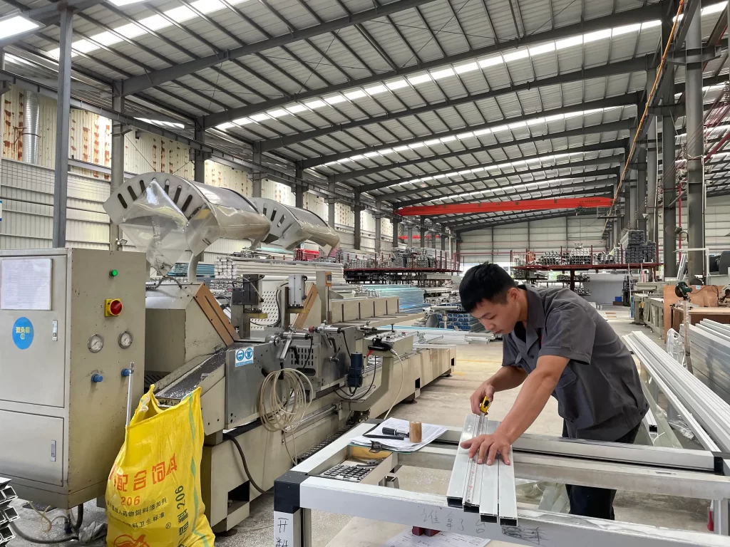 Factory worker performs precision measurement on cleanroom door components using industrial machinery, ensuring quality control in a manufacturing facility.