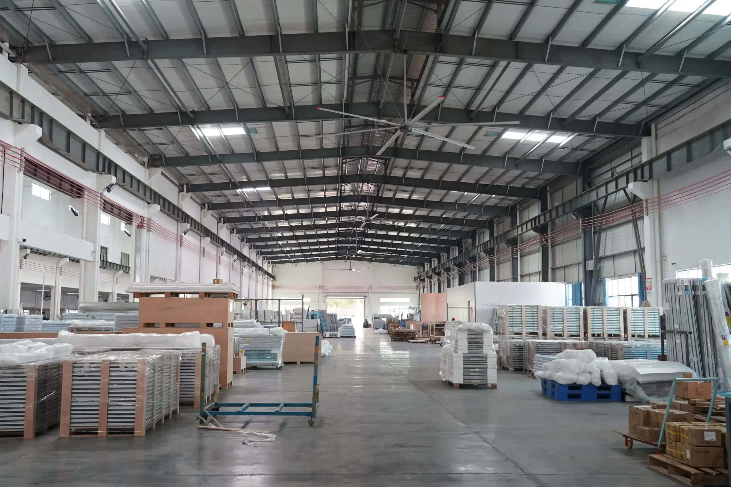 Spacious aluminum profile manufacturing warehouse with industrial ceiling fans, neatly stacked packaged finished materials and wooden pallets, a tidy production storage space.