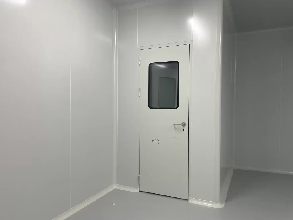 A cleanroom accessory: white industrial cleanroom door with glass view window, in an empty food and beverage cleanroom with smooth wall panels.