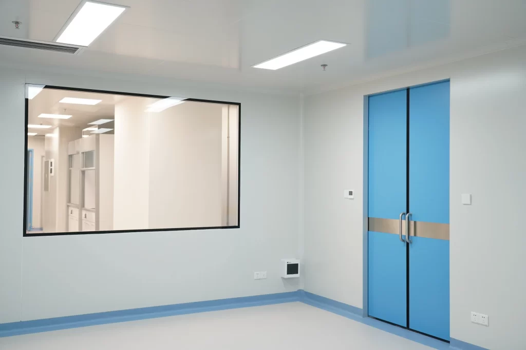 Pharmaceutical cleanroom interior featuring a large observation window and blue sliding door, with smooth walls and ceiling fixtures.