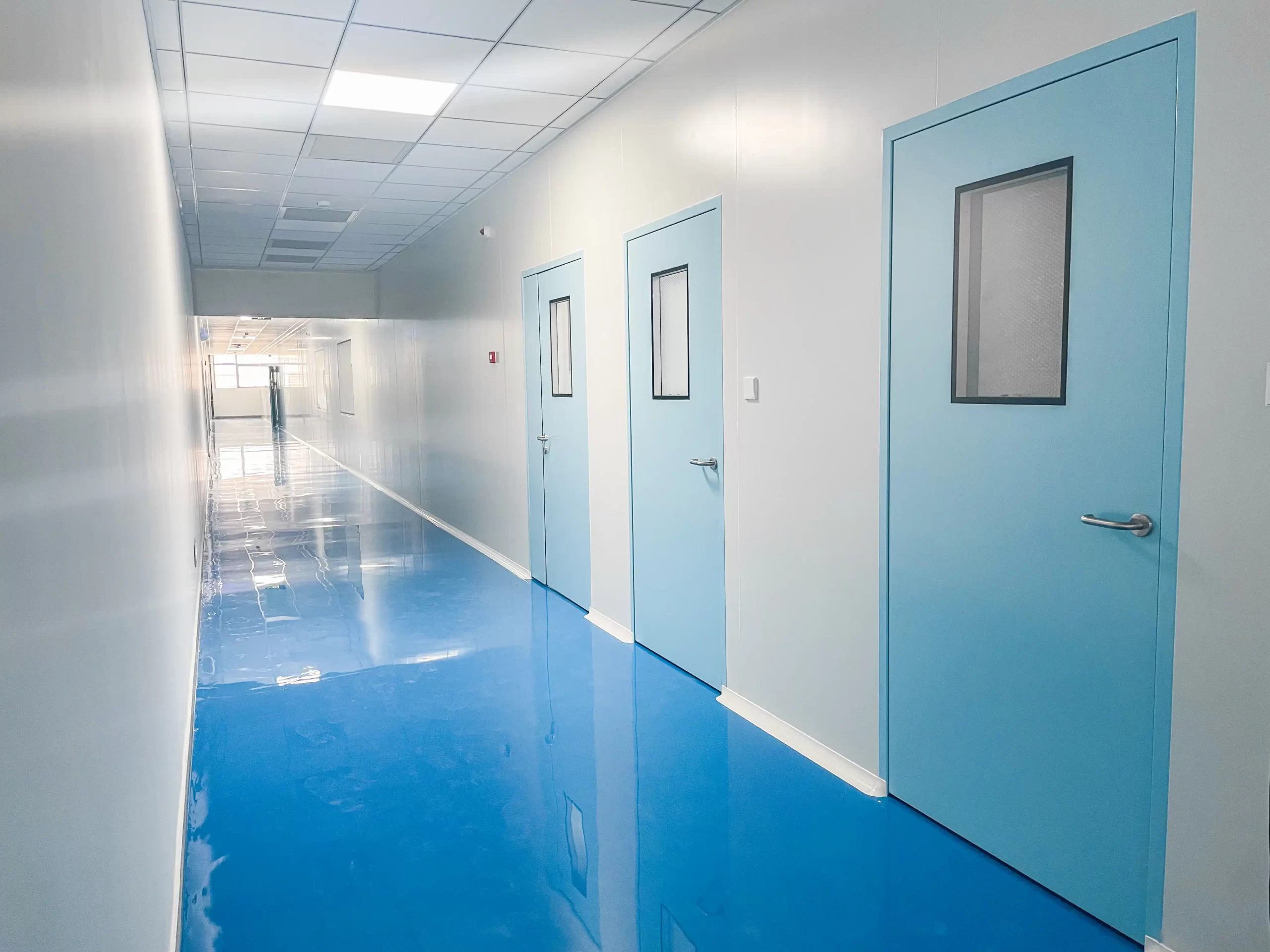 Sterile pharmaceutical cleanroom corridor with light blue GMP-compliant doors, smooth blue flooring, and bright white paneled walls.