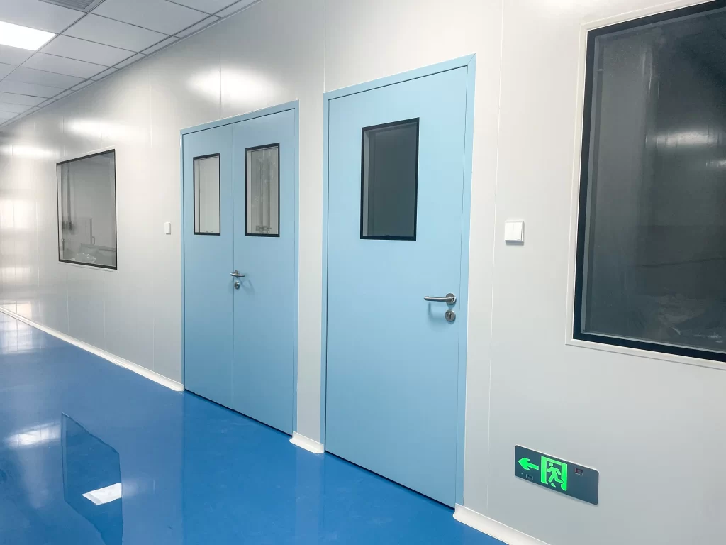 Corridor view of a cleanroom facility with light blue single and double laboratory doors, white walls, blue floor, and safety exit sign.
