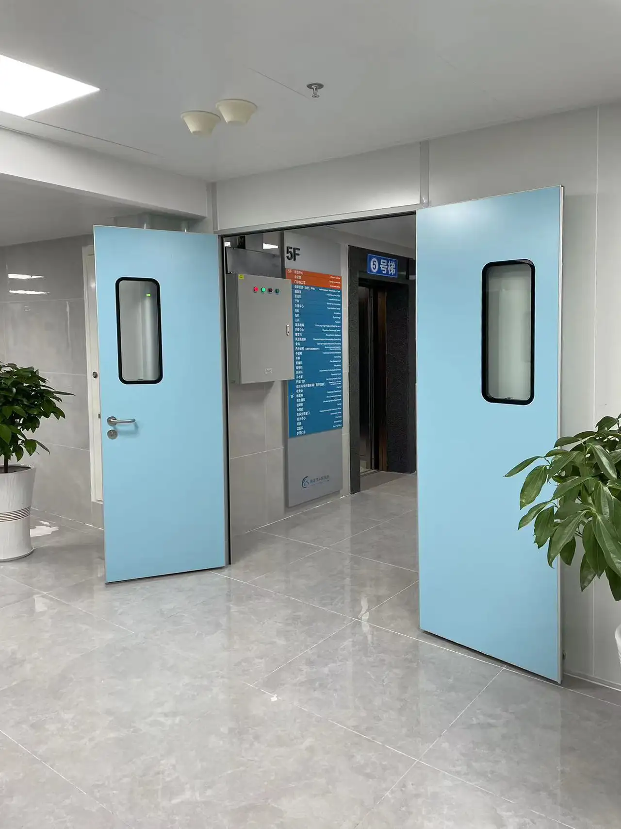 Open light blue hospital double doors revealing a polished tile corridor, elevator lobby, and floor directory sign.