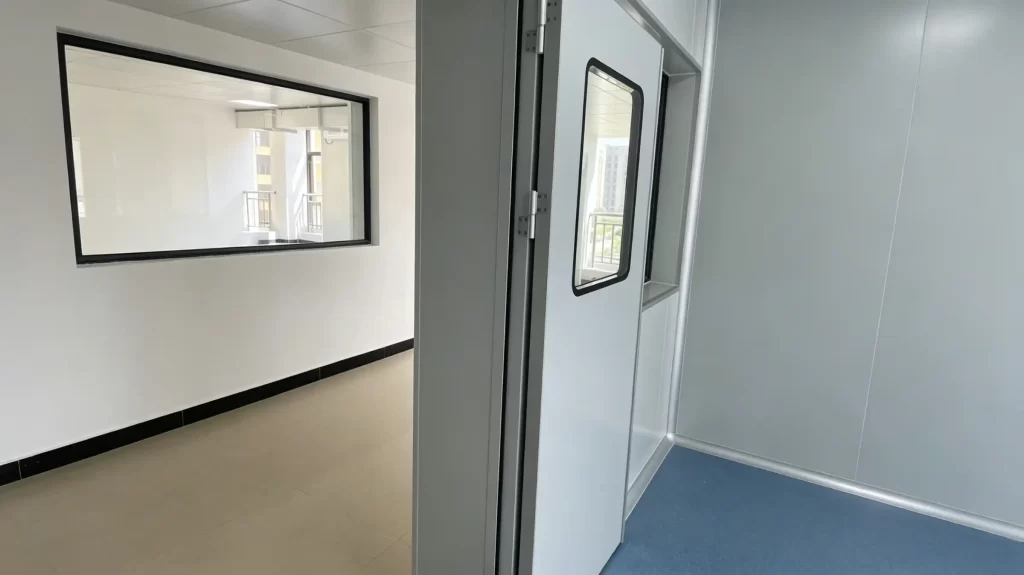 Modern cleanroom corridor featuring large observation windows with black frames, white walls, beige flooring with black baseboards, and glass doors leading to controlled environment areas