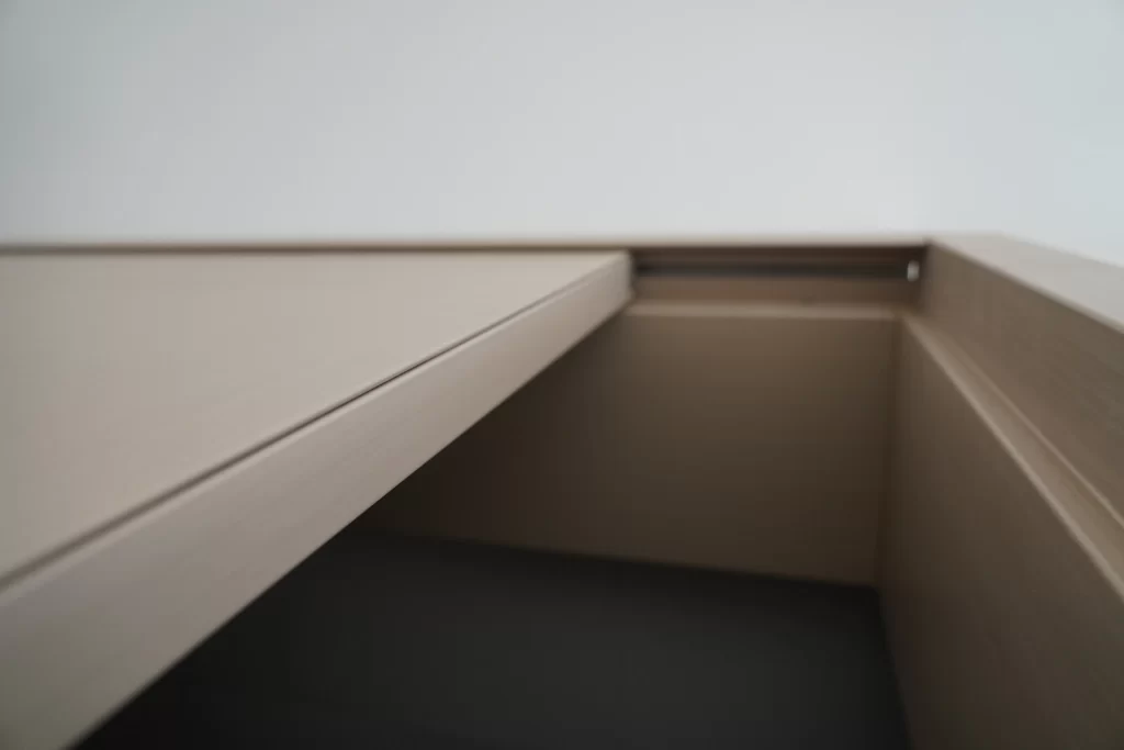 Close-up view of a light wood-grain door frame or furniture edge detail against a white wall and dark floor.