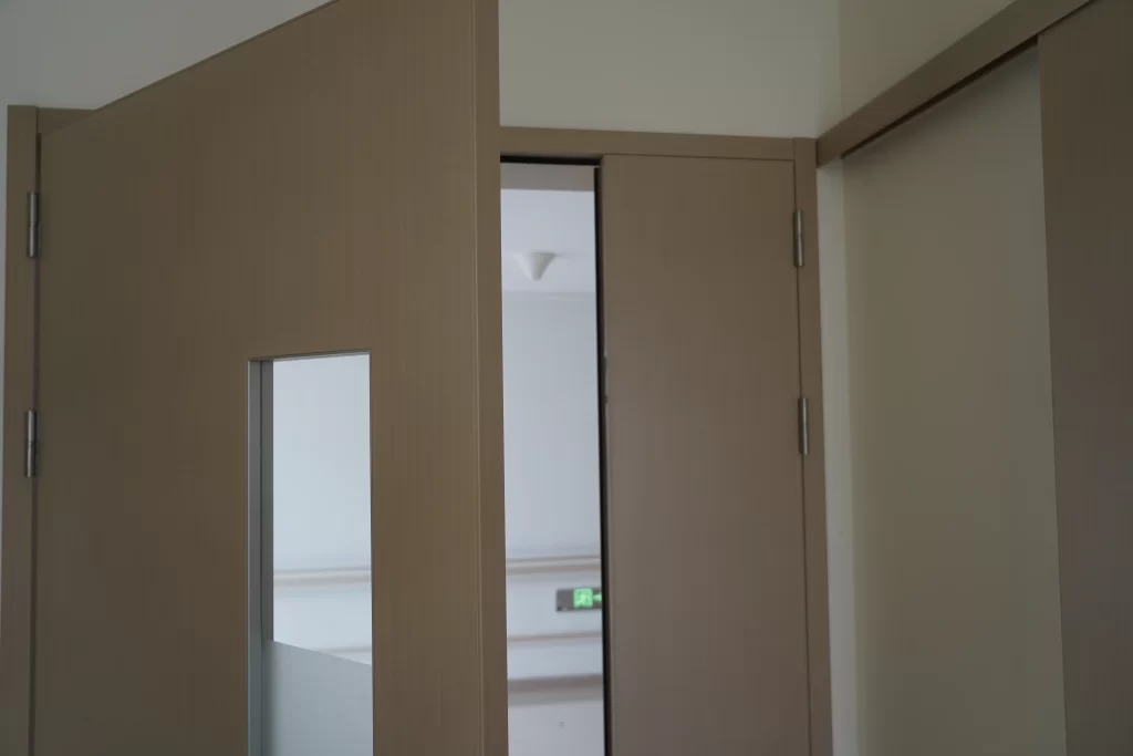 Beige hospital corridor doors with vertical vision panels, one slightly open revealing a hallway and a green safety exit sign.