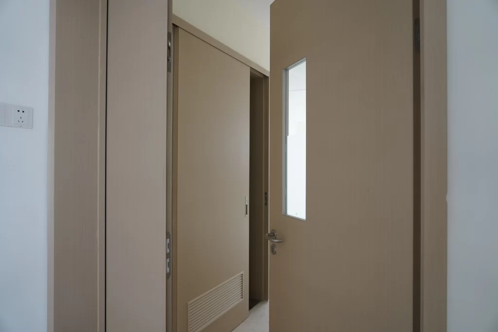 A beige hospital patient room door featuring a vertical frosted glass vision panel, silver handle, and bottom ventilation louver.