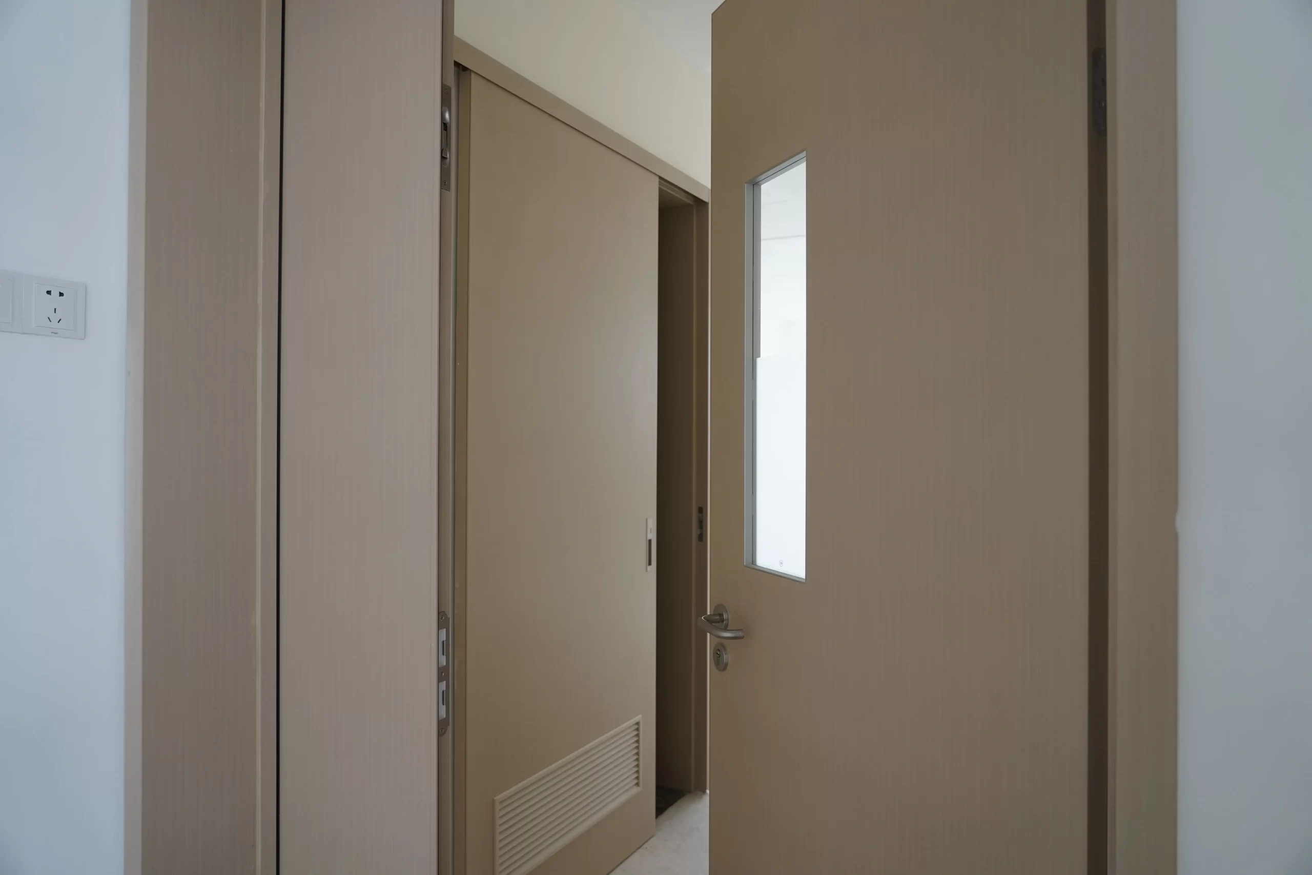 A beige hospital patient room door featuring a vertical frosted glass vision panel, silver handle, and bottom ventilation louver.
