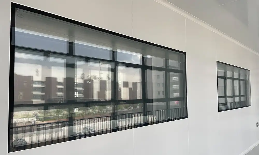 Large black-framed observation windows on a white wall near a pharmaceutical cleanroom door, showing exterior buildings.