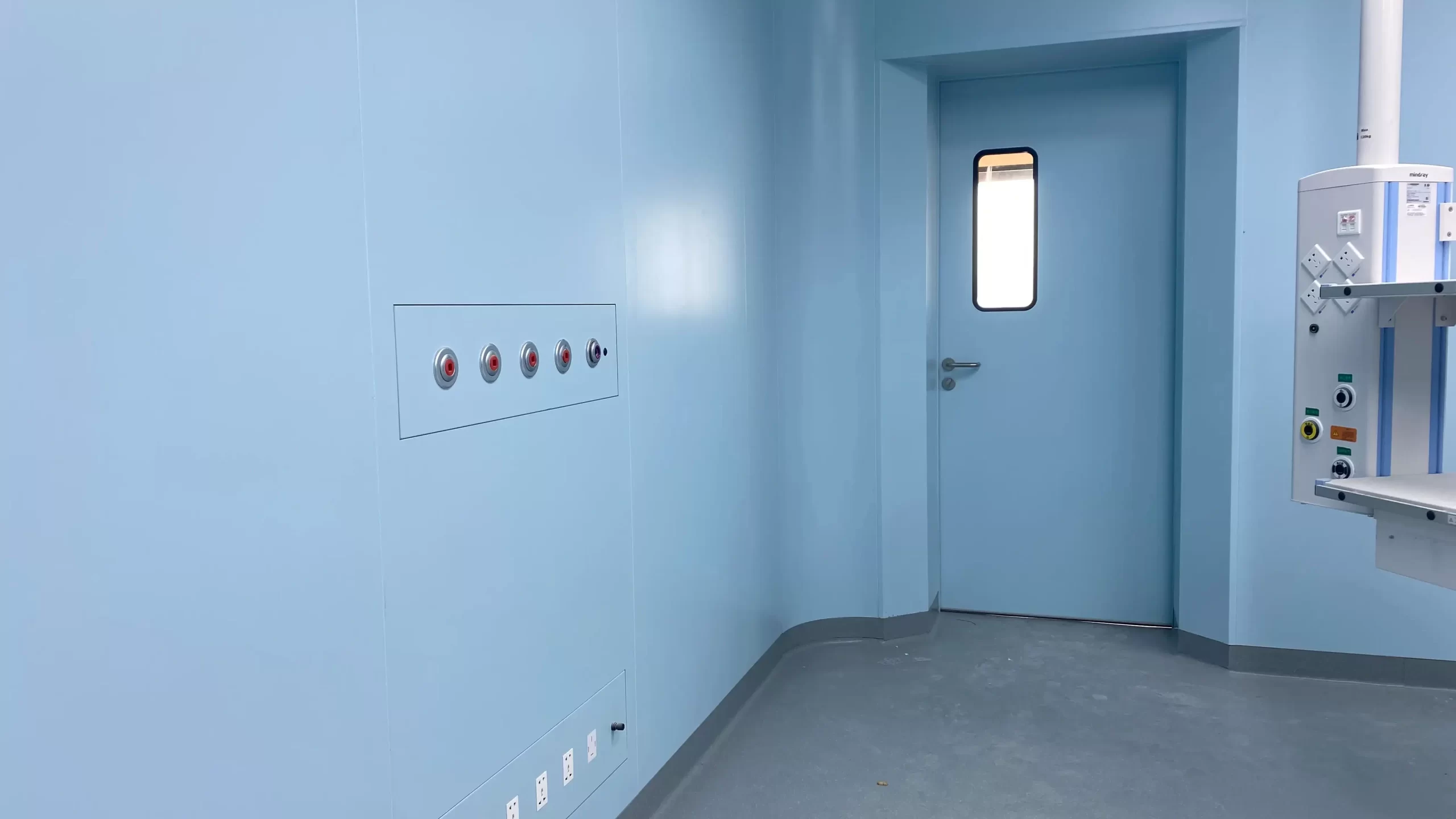 Cleanroom corner featuring a single-leaf hinged door with a vertical window, wall control panel, and a medical pendant.