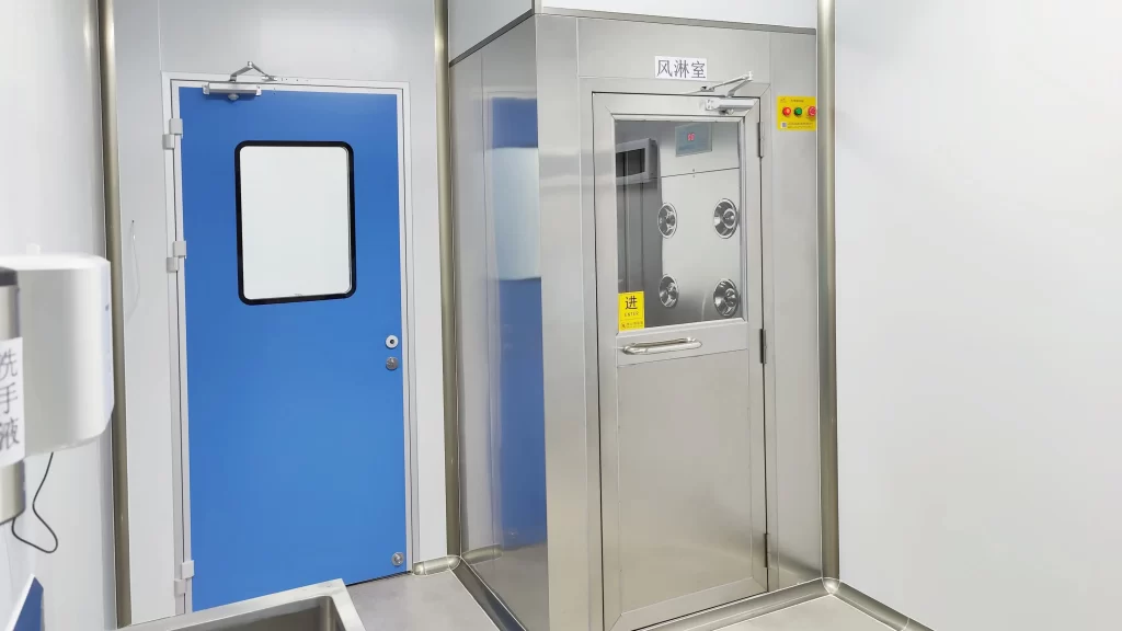 Biotech cleanroom gowning area featuring Cleanroom pass door paired with stainless air shower interlock chamber, flush vision window, concealed hinges, compliant ISO 7 gowning prep zone.