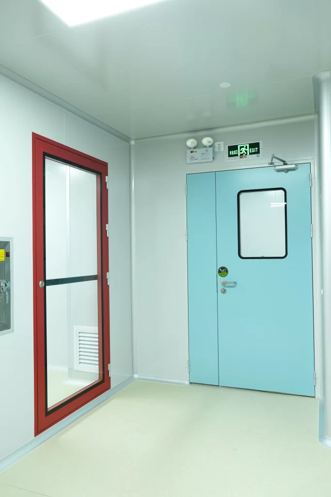 Cleanroom interior showing a red-framed glass door and a blue double fire exit door with an illuminated green EXIT sign.