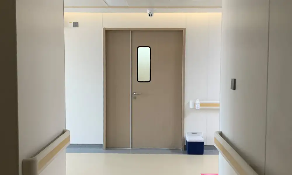 Bright hospital corridor with beige wood-grain double swing hygienic door, vertical observation window, wall handrails, light neutral medical interior and smooth sterile flooring
