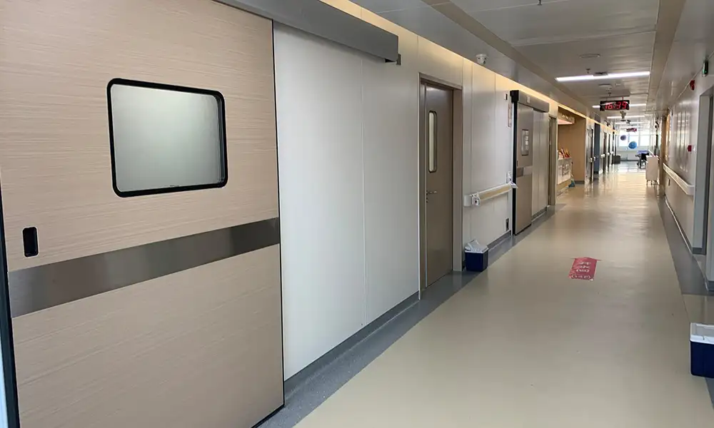 Long hospital ward corridor with wood-grain medical self-closing sliding door and swing patient room doors, hygienic controlled healthcare environment.