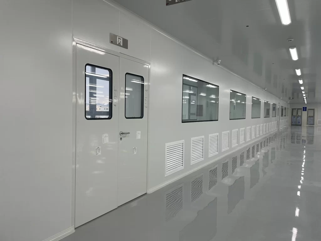 A long cleanroom corridor with white walls, ventilation grilles, and stainless steel double doors with viewing windows.