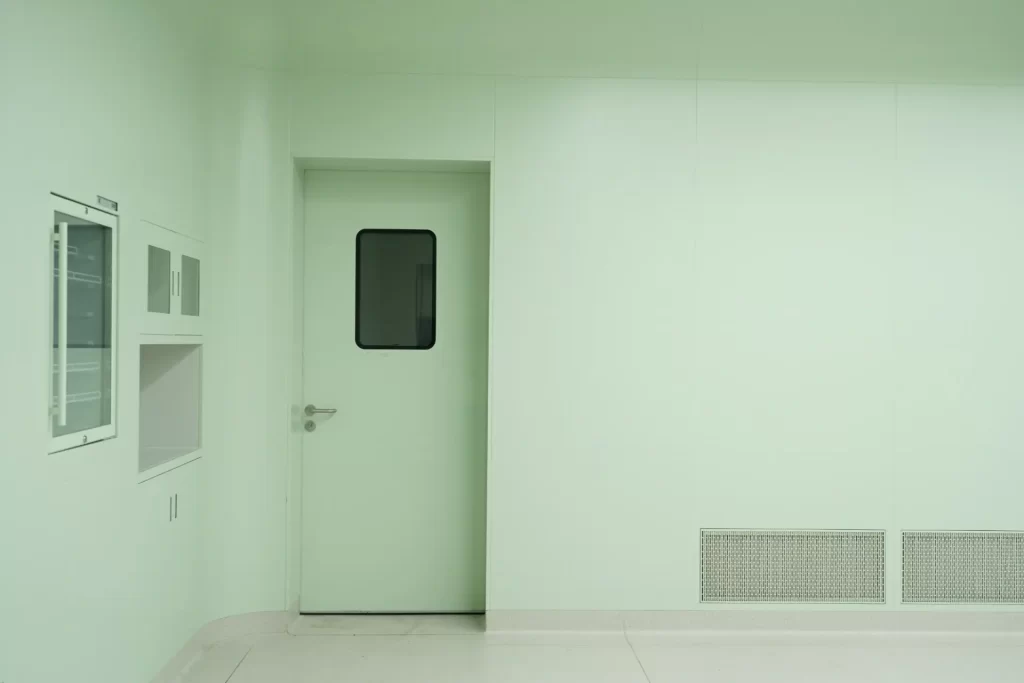 E-ZONG single swing cleanroom door featuring a glass vision panel, flush-mounted wall system, pass-through box, and integrated return air grill in a green-tinted pharmaceutical cleanroom environment.
