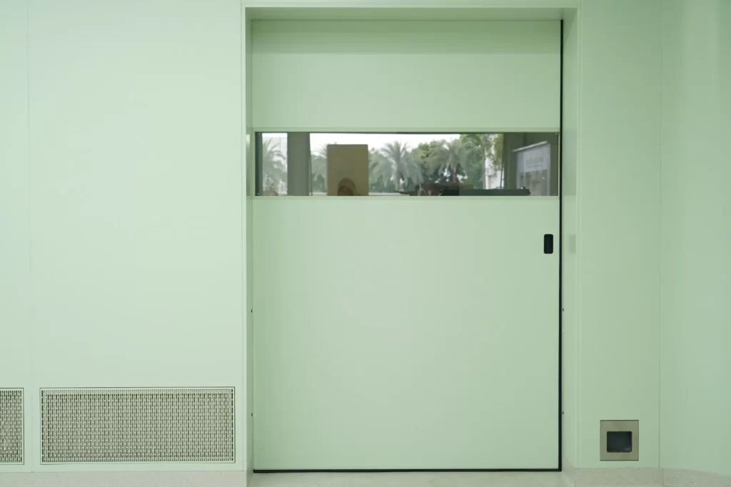 Light green automatic sliding cleanroom door with a horizontal glass observation window fitted in a pharmaceutical facility wall.