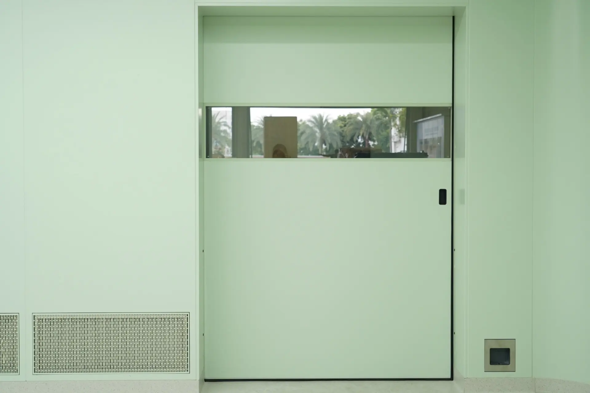 Light green automatic sliding cleanroom door with a horizontal glass observation window fitted in a pharmaceutical facility wall.