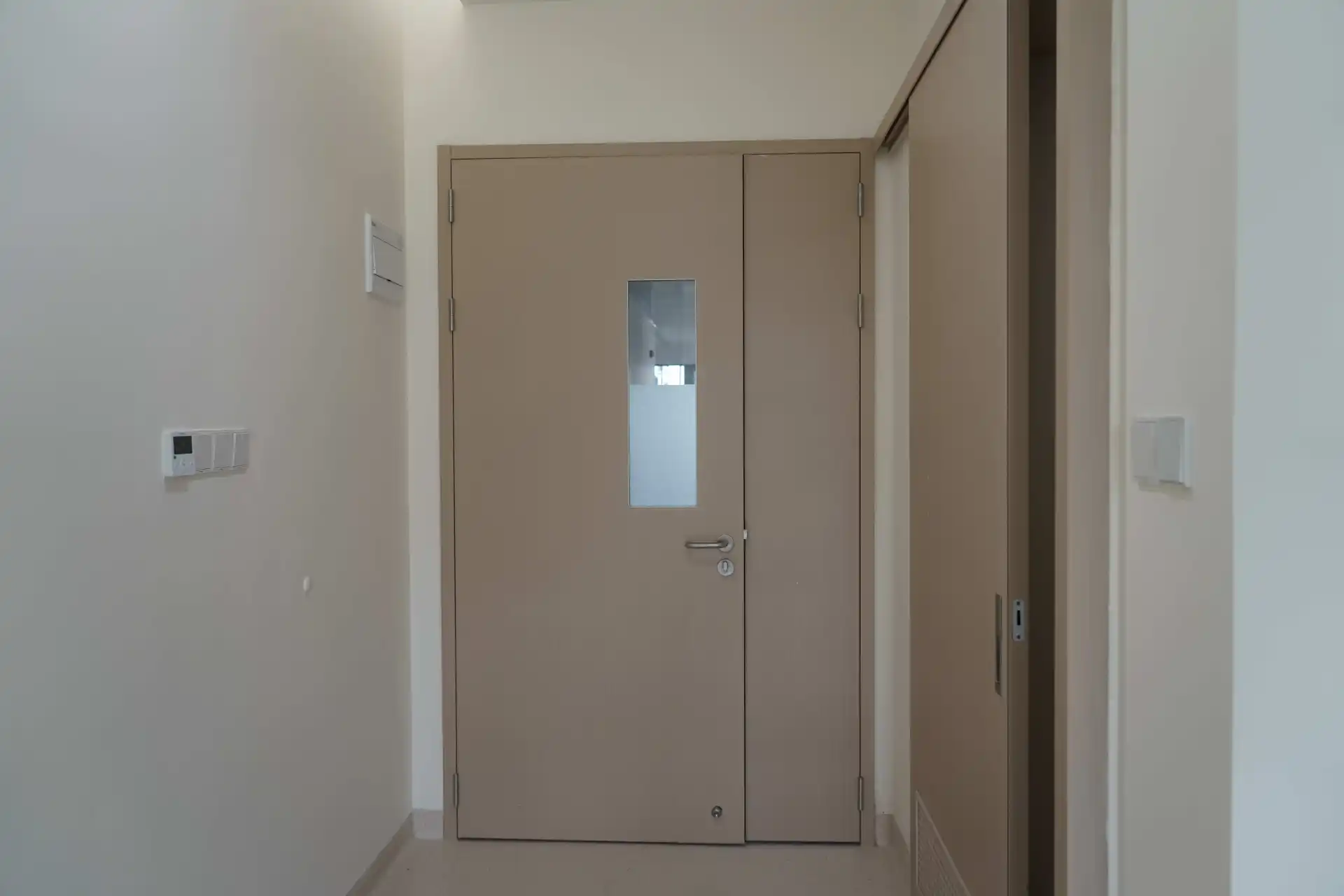 A beige unequal double swing door (mother-child door) with a glass vision window and lever handle installed in a hospital cleanroom wall.