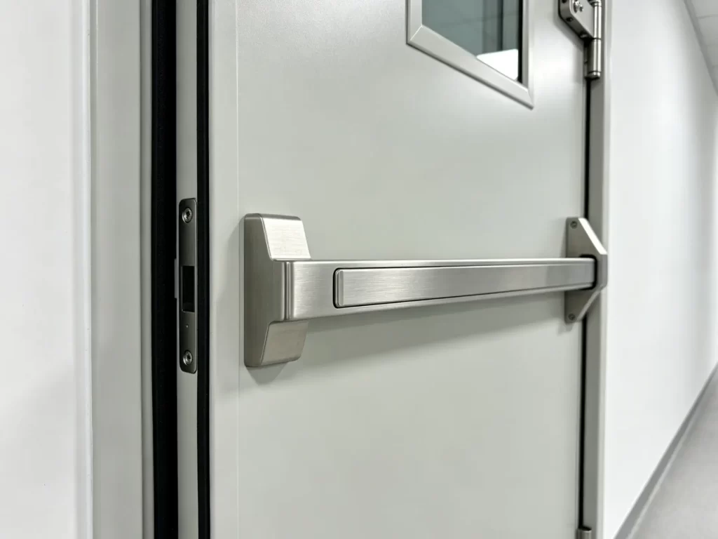 Detailed view of a stainless steel panic exit push bar and mortise latch installed on a flush cleanroom door.