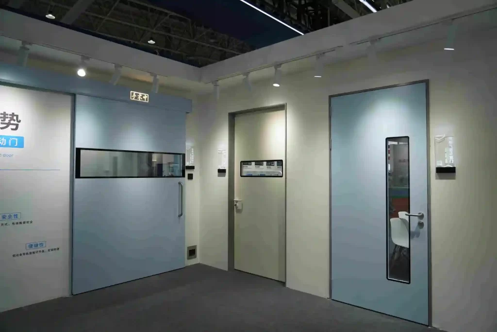 A display at a trade show featuring three types of medical-grade hospital doors: a large light-blue sliding door for an operating room (left), a beige single swing door with a window (center), and a blue single swing door with a narrow window (right)