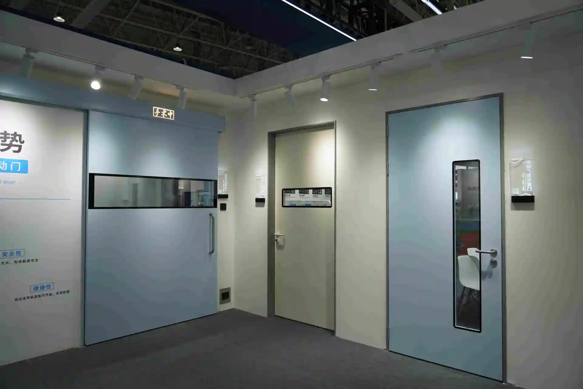 A display at a trade show featuring three types of medical-grade hospital doors: a large light-blue sliding door for an operating room (left), a beige single swing door with a window (center), and a blue single swing door with a narrow window (right)