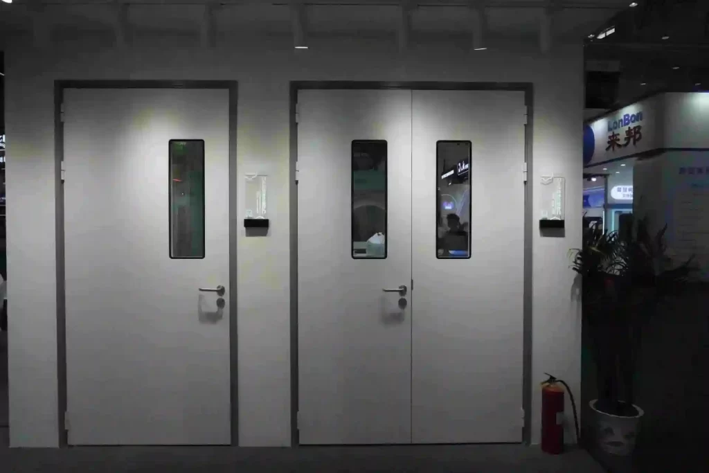 A front view of two clean, white, single-leaf swing hospital doors with rectangular vision panels and stainless steel lever handles, installed within a wall at an industry exhibition.